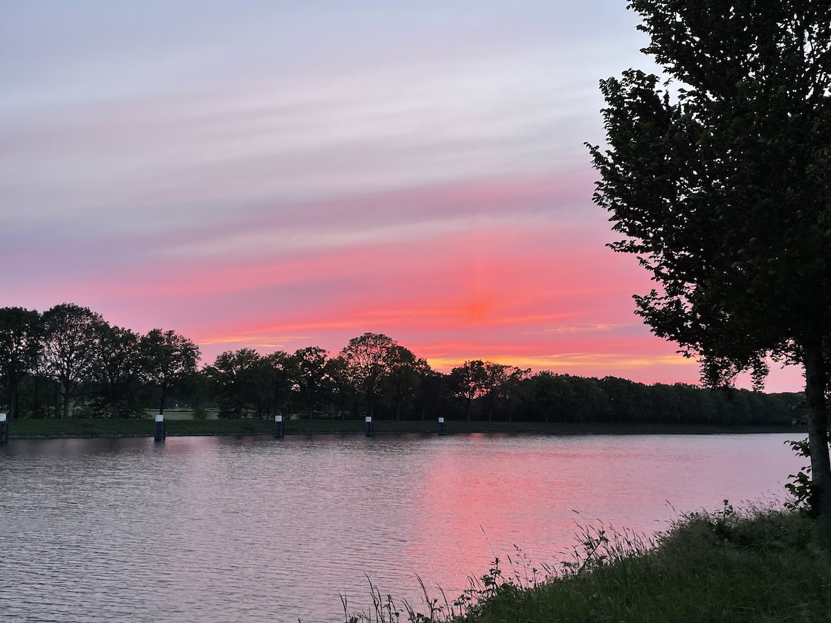 Foto van prachtig avondrood, weerspiegeld in een kanaal. Met donkere contouren van bomen en grassen die tegen de lucht en het kanaal afsteken.